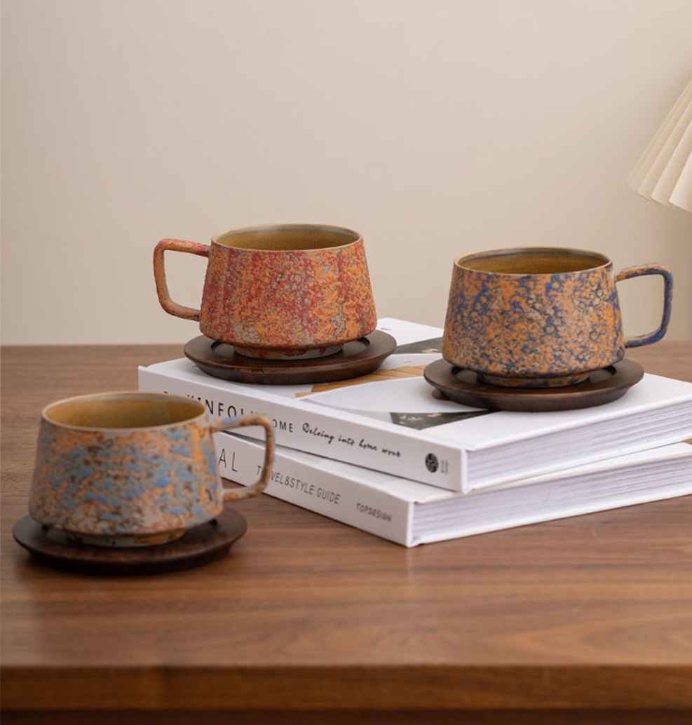 Three mottled cups in red, ochre, and blue-grey arranged on books and a wooden table